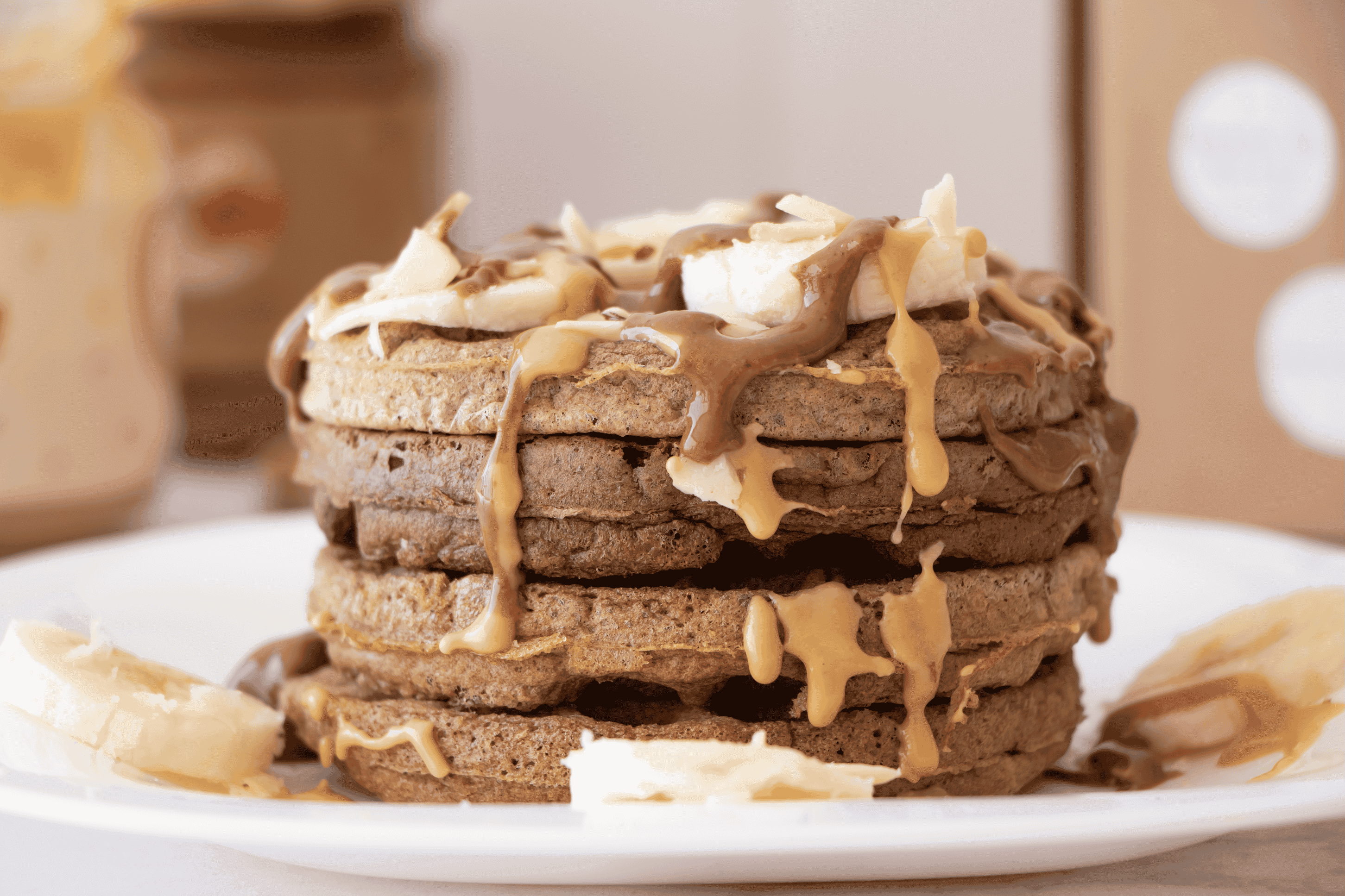 Torre de waffles con plátano y crema de cacahuate cayendo desde una cuchara, junto a los frascos de untables
